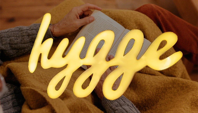 Ilustración del concepto Hygge: persona leyendo cómodamente en un sofá con manta y bebida caliente.