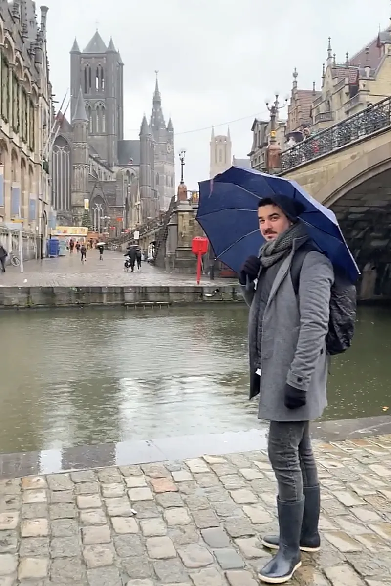 Álex con paraguas y botas de agua en el muelle de Graslei de Gante, junto al puente de San Miguel, con la iglesia de San Nicolás y el Belfort al fondo