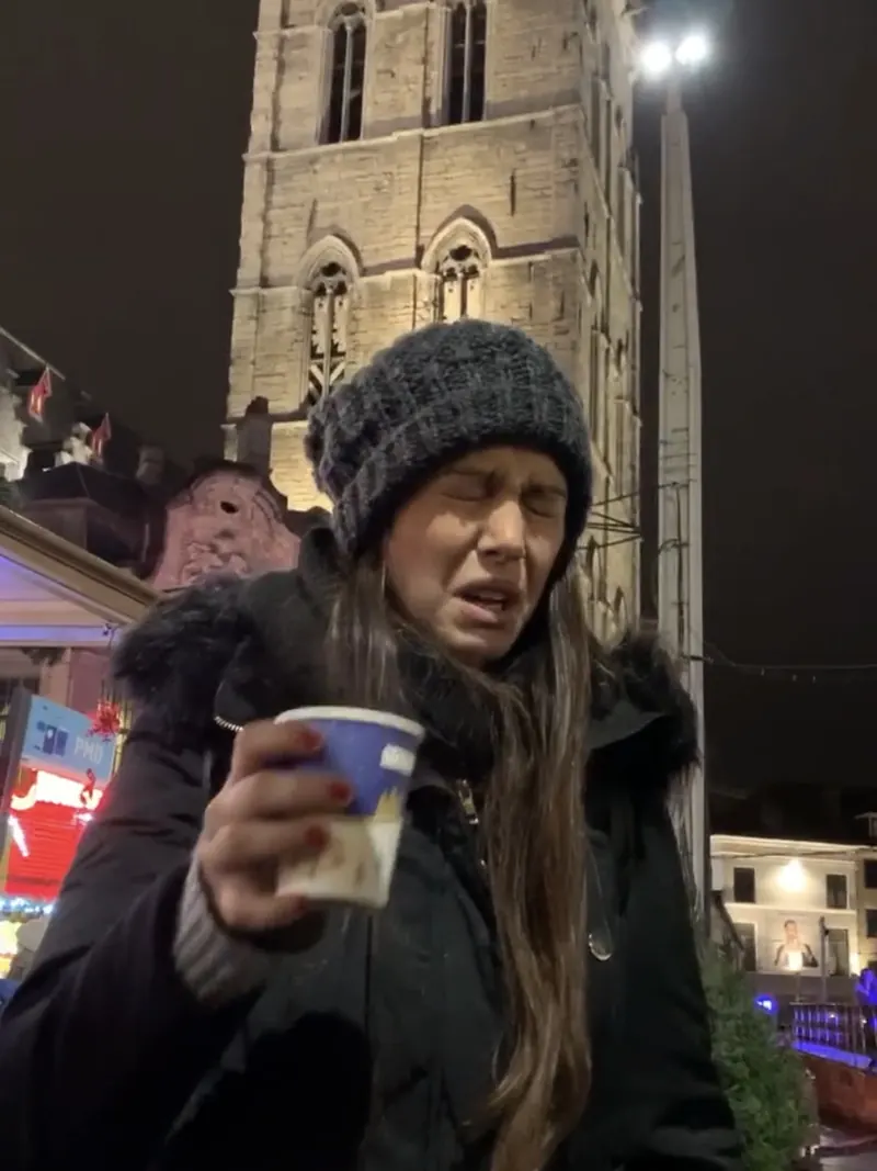 Inma haciendo una mueca tras probar vino caliente en el mercado navideño de Gante, de noche, con una torre iluminada detrás