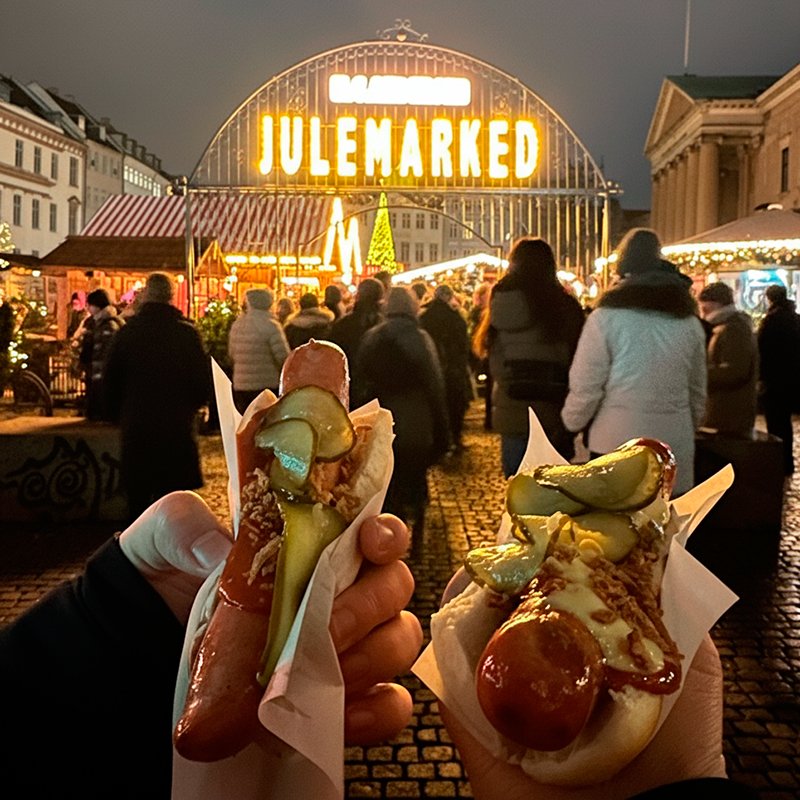 Dos 'pølser' daneses con pepinillos y salsa, sostenidos en primer plano en un mercadillo navideño de Copenhague iluminado.