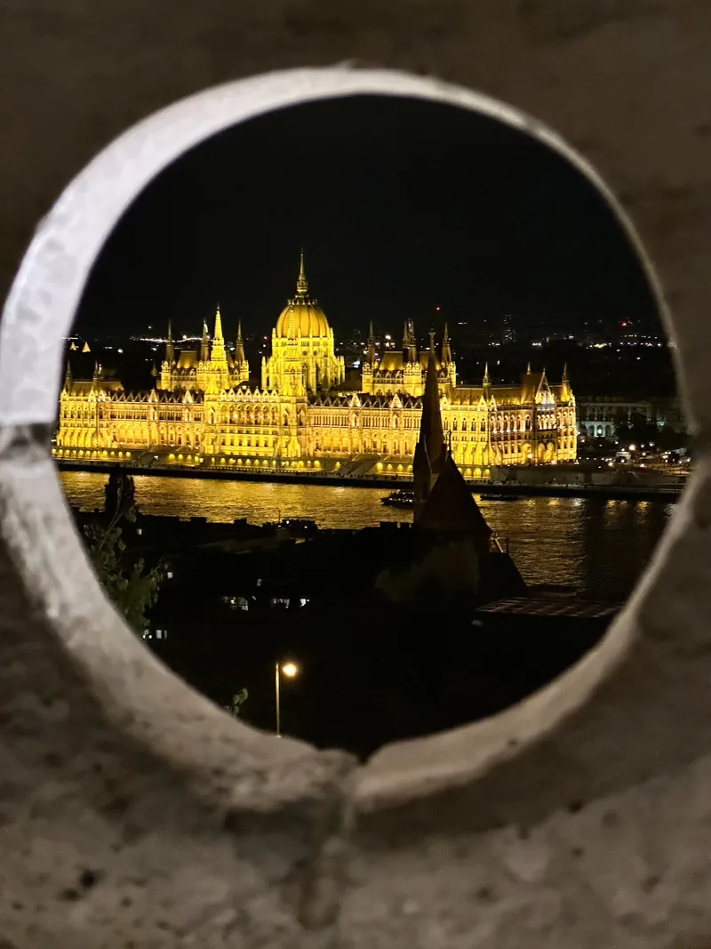 El Parlamento de Budapest iluminado de noche, visto a través de un hueco redondo del muro del Bastión de los Pescadores