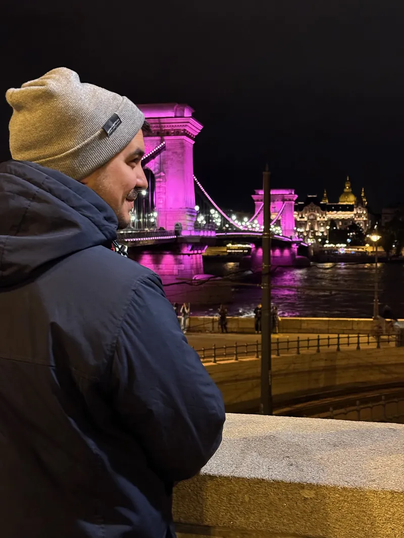 Álex con gorro y abrigo frente al Puente de las Cadenas iluminado de rosa, de noche en Budapest